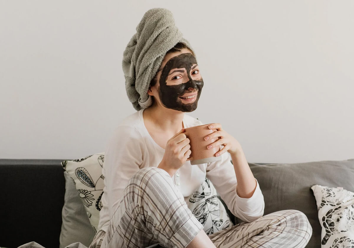 Image of woman wearing a face mask. She is sitting on her couching and drinking tea.
