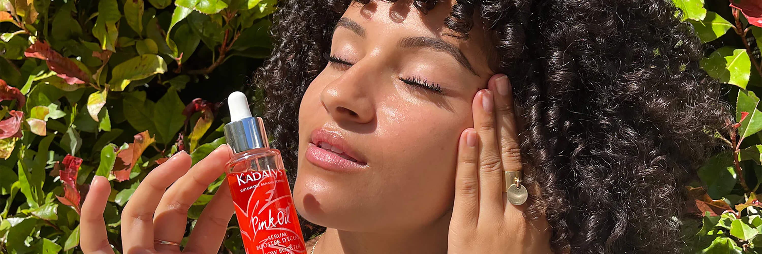 Black woman applying pink banana oil to her skin in front of green shrubbery | Kadalys natural skincare products to help dullness, uneven tone, and hyperpigmentation