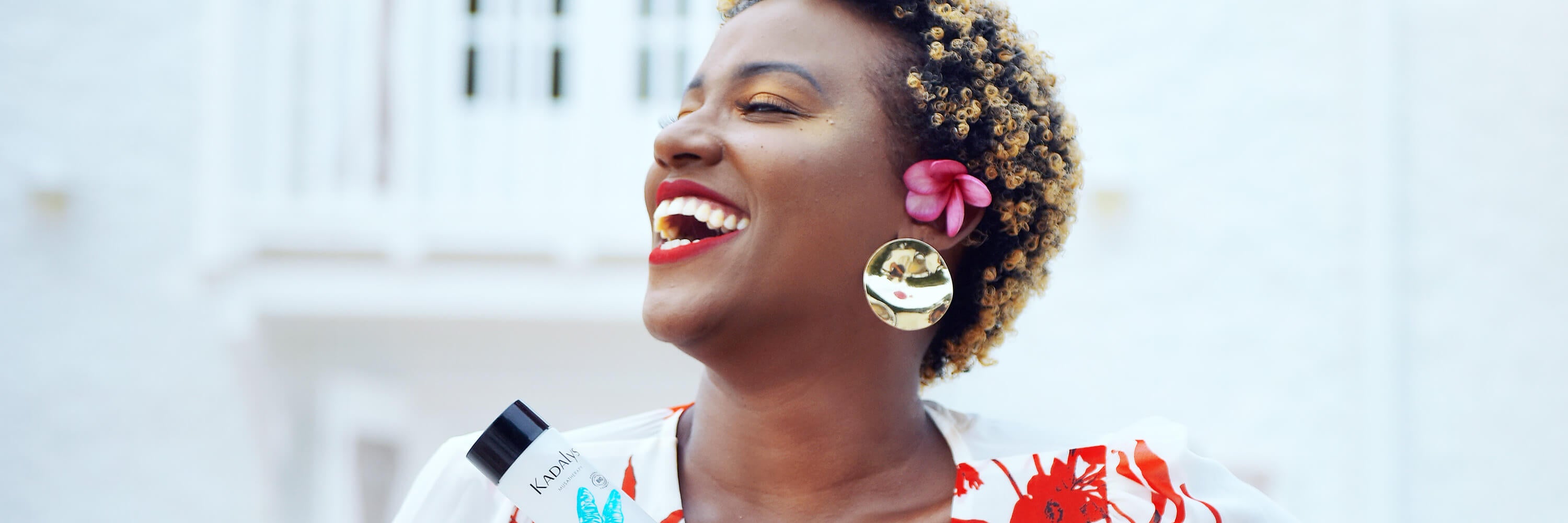 Black Caribbean woman smiling broadly in front of white building and holding a Kadalys product | Kadalys natural vegan skincare products