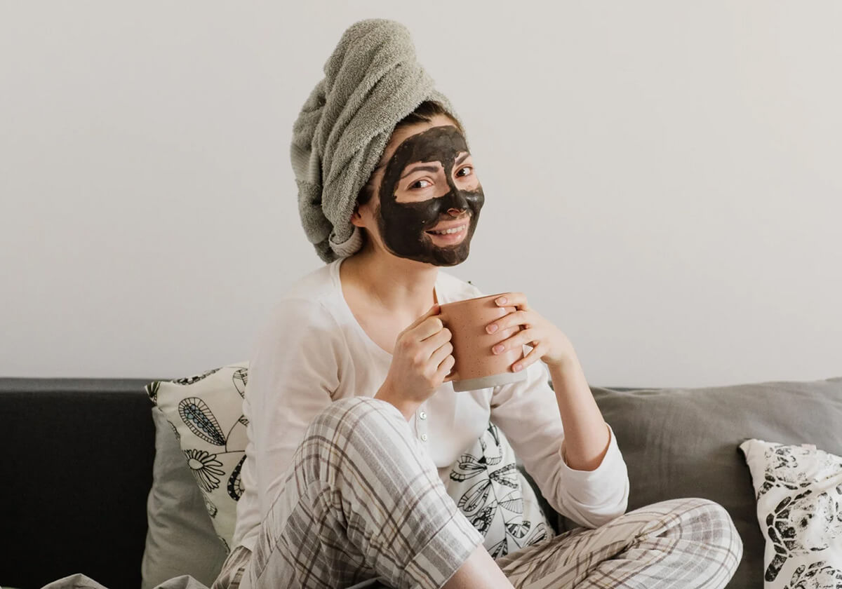 Image of woman wearing a face mask.  She is sitting on her couching and drinking tea.